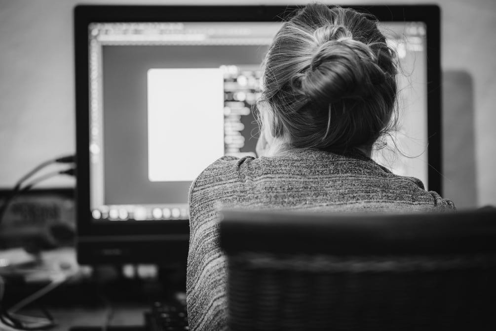 Black and white rear view of a woman with her hair in a bun working at a computer with a large monitor