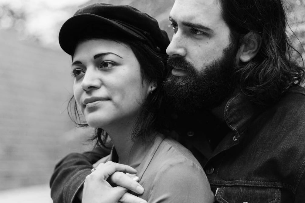 Black and white portrait of a bearded man embracing a woman from behind as they both look thoughtfully into the distance